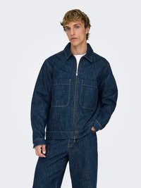 Mørkeblå denimjakke med glidelås foran, to brystlommer og spiss krage. Brukes med matchende mørkeblå bukser. Jevn tekstur.