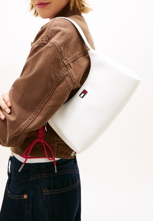 Γυναίκα που φοράει καφέ μπουφάν και σκούρα τζιν κρατάει μια λευκή τσάντα ώμου με ένα μικρό ορθογώνιο λογότυπο σε λευκό φόντο.