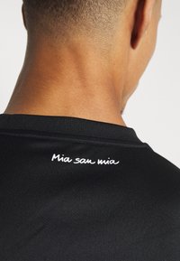 Maglietta atletica nera con una texture liscia e il dettaglio del testo "Mia san mia" in bianco sul colletto posteriore.