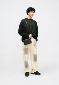 Melnas oversize džemperis ar grafisku apdruku, krēmkrāsas bikses ar tumšiem plankumiem un melni sporta apavi. Mazs melns plecu soma.