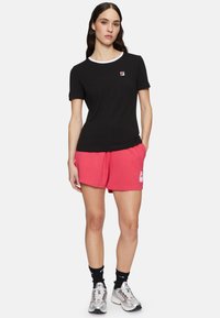 Camiseta negra de cuello redondo con ribete blanco, combinada con pantalones cortos rosas que presentan un logo. El look se completa con zapatillas plateadas y calcetines negros.