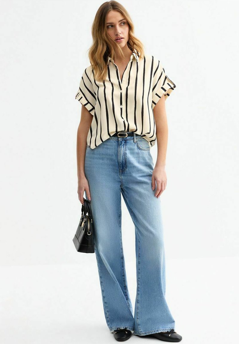 Blouse à rayures à manches courtes en crème et noir, associée à un jean large bleu clair. Tient un petit sac à main noir, chaussée de chaussures noires.