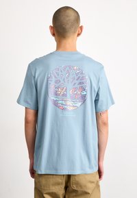 Camiseta de algodón de color azul claro que presenta un gráfico circular de un árbol con detalles e colores intrincados. El texto debajo dice "Ilustrado por Austin Rostborough."