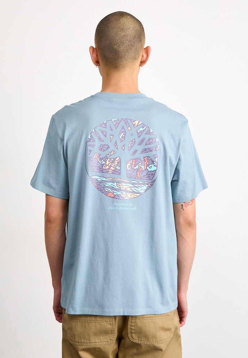 Camiseta de algodón de color azul claro que presenta un gráfico circular de un árbol con detalles e colores intrincados. El texto debajo dice "Ilustrado por Austin Rostborough."