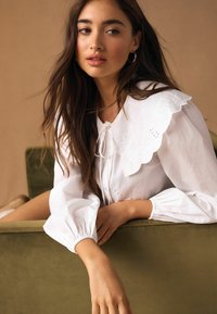 Fehér blúz hullámos szegélyű gallérral és virágos hímzéssel, fodros ballonujjakkal és elöl megkötős záródással, semleges háttér előtt.