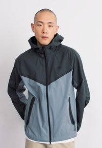 Windbreaker jacket featuring a dark gray upper section and light gray lower section, with a zip closure and two side pockets.