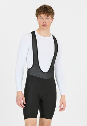 Homme portant un cuissard de cyclisme noir sur un maillot blanc à manches longues, debout devant un fond clair uni.