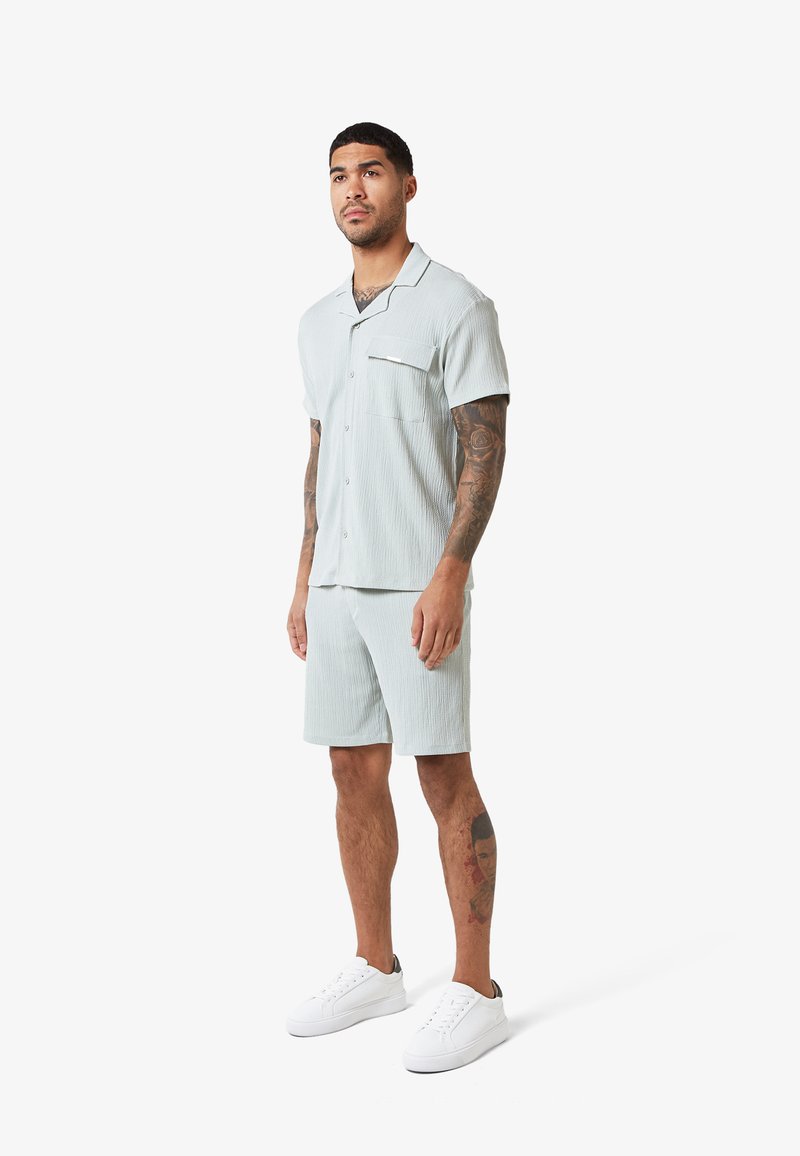 Άνδρας με τατουάζ που φοράει ανοιχτό μπλε πουκάμισο με κοντό μανίκι και κουμπιά, καθώς και αντίστοιχα σορτς, με λευκά αθλητικά παπούτσια, στέκεται προς τα εμπρός.