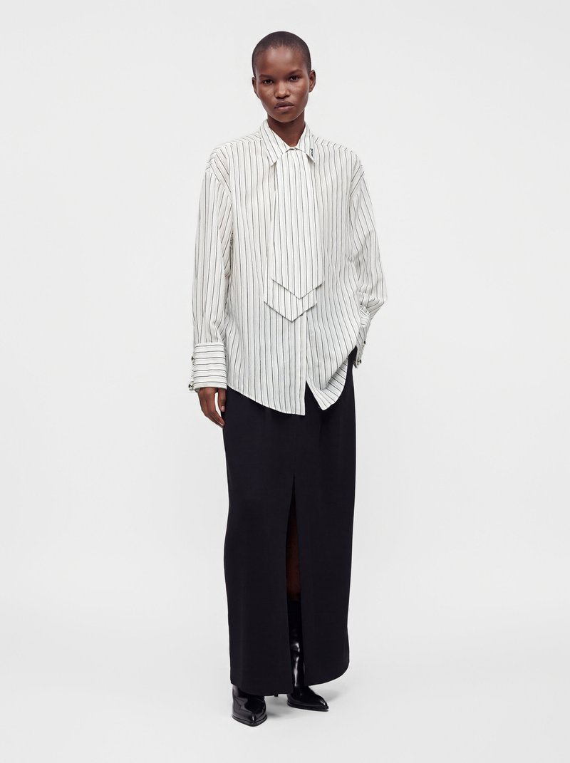 Personne debout portant une chemise blanche à fines rayures oversize avec de larges poignets, une longue jupe noire fendue sur le devant, et des bottes noires pointues.