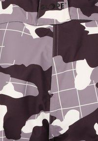 Lila och vit kamouflage-mönstrad tyg med slät textur. Har en vertikal söm och en tryckt logotyp ovanför. Rutnätslinjer är synliga i designen.