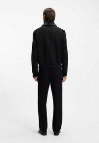 Mann, der mit dem Rücken zur Kamera steht, trägt eine schwarze Jacke mit Pelzkragen, schwarze Hose und schwarze Schuhe vor weißem Hintergrund.