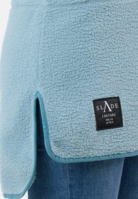 Indumento in pile di colore azzurro chiaro con superficie testurizzata, spacco laterale e un'etichetta nera con la scritta "SLADE COUTURE" sul bordo inferiore.