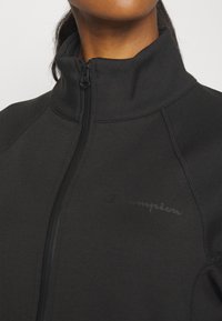 Svart zip-up jacka i mjuk tyg, med hög krage, figurnära design och en subtil Champion-logotyp på framsidan.