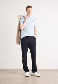Polo de culoare albastru deschis cu detalii închise, pantaloni navy și adidași albi; model ținând o jachetă bej pe umărul unuia, stând pe un fundal neutru.