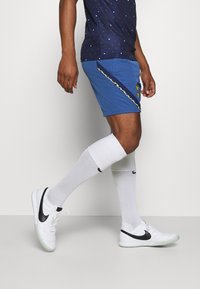 Pantaloni sportivi blu con un motivo testurizzato, dettagli gialli e bordi bianchi. Abbinati a un logo Nike nero e calze bianche fino al ginocchio e scarpe.