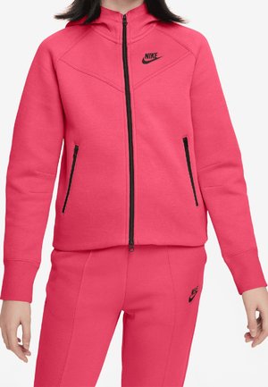 Donna che indossa una tuta Nike rosa acceso con cerniera nera e logo, composta da una giacca con cerniera e pantaloni abbinati.