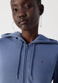 Haut à fermeture éclair bleu fabriqué en tissu doux, avec une capuche, des cordons de serrage et un petit logo brodé sur la poitrine. Boucles d'oreilles en argent visibles.