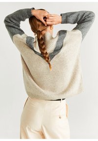 Pull en tricot beige et gris avec une texture côtelée, présentant un motif en forme de V dans le dos. Associé à un pantalon de couleur claire.