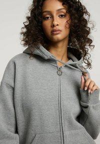 Szara hoodie na zamek, wykonana z miękkiego materiału, z kapturem na sznurku, przednimi kieszeniami i ściągaczami; posiada srebrną końcówkę zamka.