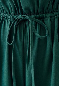 Tessuto verde testurizzato con una fascia elastica in vita, con un laccio annodato. Superficie liscia con morbide pieghe nel materiale.