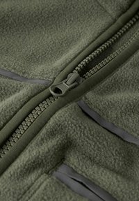 Primer plano de una chaqueta de forro polar verde con la cremallera parcialmente desabrochada y costuras visibles en la tela texturizada.