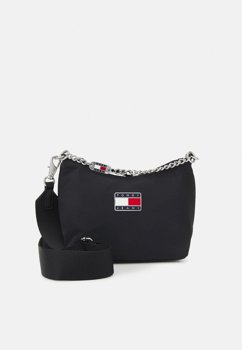 Sac d'épaule Tommy Jeans noir avec détail chaîne argentée, patch logo rectangulaire et sangle en tissu réglable sur fond uni.