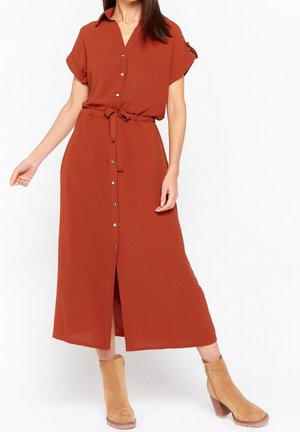 Vrouw in een midi-jurk met knopen in roestoranje, korte omgeslagen mouwen, een taillekoord en beige suède enkellaarsjes met blokhakken.