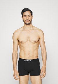 Mustat puuvillaboksereet, joissa on valkoinen "Levi's"-logo elastisessa vyötärönauhassa, istuva malli, esitelty paljaalla vartalolla yksinkertaista taustaa vasten.