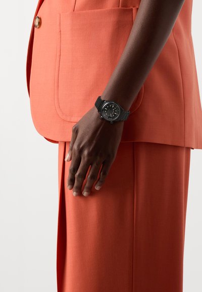 Black wristwatch with a round face, rubber strap, set against an orange textured suit. Hand rests on the suit’s pocket.