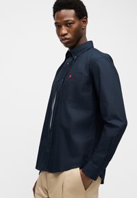 Mężczyzna w ciemnogranatowej koszuli zapinanej na guziki z kieszenią na piersi i beżowych spodniach, stojący z jedną ręką w kieszeni na jednolitym tle.