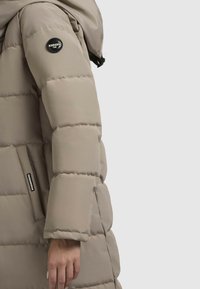 Beige színű pufikabát, kapucnival, vízszintes steppeléssel, kerek logo folttal a karon és oldalzsebbel, amelyen egy díszcímke található.