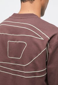 Osoba s fialovým svetrom, na zadnej strane s bielymi geometrickými švovými líniami, z pohľadu zozadu a mierne otočená doprava.