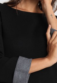 Top in maglia nera con scollatura ampia e polsini a coste grigi. Il tessuto sembra morbido e ha una texture liscia. Un piccolo tatuaggio è visibile sul polso.