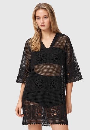 Jeune femme portant une robe en filet crocheté noire avec des motifs "ASA", debout devant un fond clair uni.