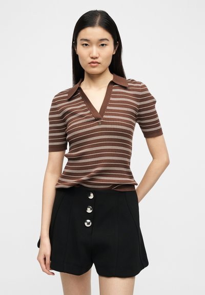 Jeune femme aux cheveux lisses et foncés portant un t-shirt à manches courtes rayé marron et blanc et un short taille haute noir à trois boutons.