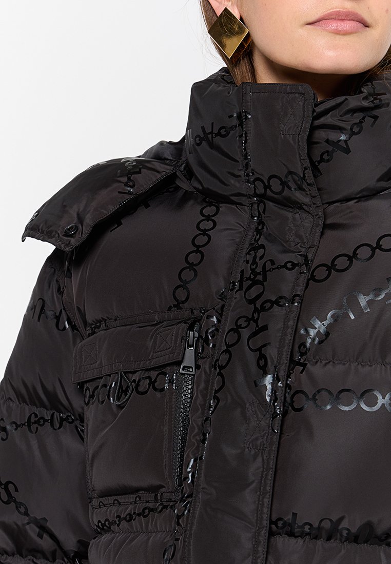 Mujer con chaqueta de plumas negra con un patrón de cadena brillante, cuello alto, bolsillo en el pecho con cremallera y grandes pendientes geométricos dorados.