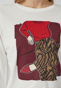 Biała koszula z długim rękawem z graficznym wzorem przedstawiającym osobę w czerwonej górze i wzorzystych brązowych spodniach, z czerwonymi ozdobnymi butami.