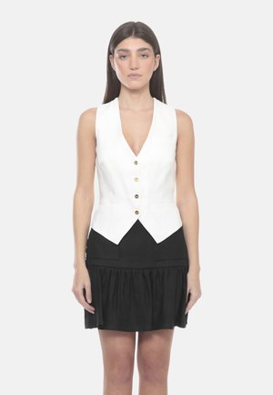 Donna con capelli lisci e scuri che indossa un gilet bianco senza maniche con bottoni e una gonna nera, in piedi davanti a uno sfondo chiaro e uniforme.