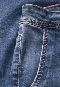 Der blaue Jeansstoff zeigt eine Nahaufnahme einer Tasche mit gelben Nähten. Die Textur ist leicht ausgebleicht und abgenutzt, was den lässigen Stil betont.