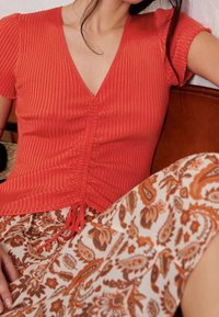 Top di corallo a coste con scollatura a V e dettagli arruffati, abbinato a una gonna crema con un motivo a paisley in arancione e marrone.