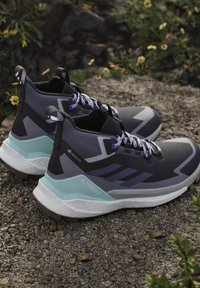 Paire de chaussures de randonnée noires et grises avec des lacets violets et des accents bleus reposant sur du gravier avec de petites fleurs jaunes à proximité.