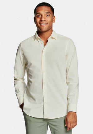 Jonge man die glimlacht, gekleed in een crèmekleurig, lang mouw shirt met knopen en lichtgrijze broek, staand tegen een effen witte achtergrond.