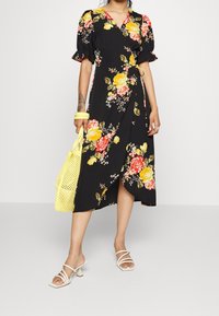 Kvinna i en svart blommig omlottklänning med gula och röda blommor, vita remsandaler, hållande en gul vävd handväska.