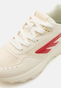 Beige idrettssko med mesh-paneler, hvite snøresnører og en rød aksentlogo. Sålen har tekstur for grep med en liten heving.