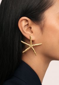 Gold textured starfish-shaped earring worn on the left ear of a person with dark hair and a dark blue garment.