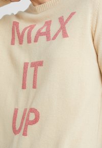 Pull en tricot beige clair avec une grande inscription en relief rose qui dit "MAX IT UP". Encolure côtelée et texture douce et lisse.
