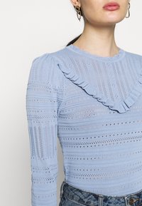 Hellblaues Langarmtop mit gerippter Textur und horizontalen Streifen, verziert mit einem Rüschensaum und offenen Strickdetails.