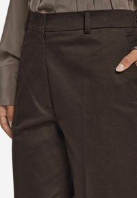 Bruna byxor med en slät, strukturerad tyg; har en rak passform och en standard midjeband. Fickor syns på sidorna.