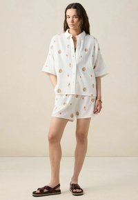 Ensemble en coton blanc comprenant une chemise à manches courtes avec boutons et un short orné de motifs de coquillage. Le tissu texturé est léger et respirant.