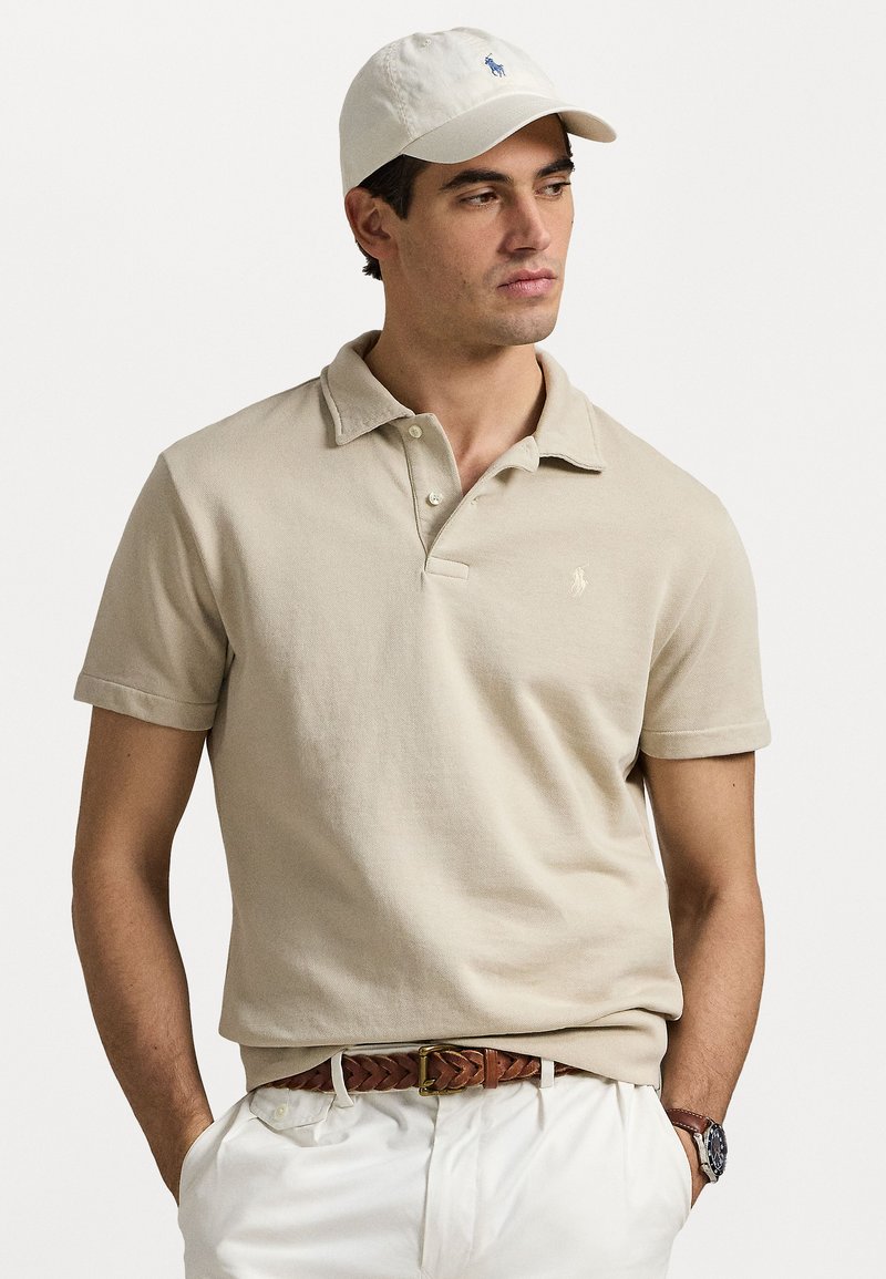 Polo beige à manches courtes et col pointu, avec un petit logo bleu. Porté avec un pantalon blanc et une casquette beige assortie.